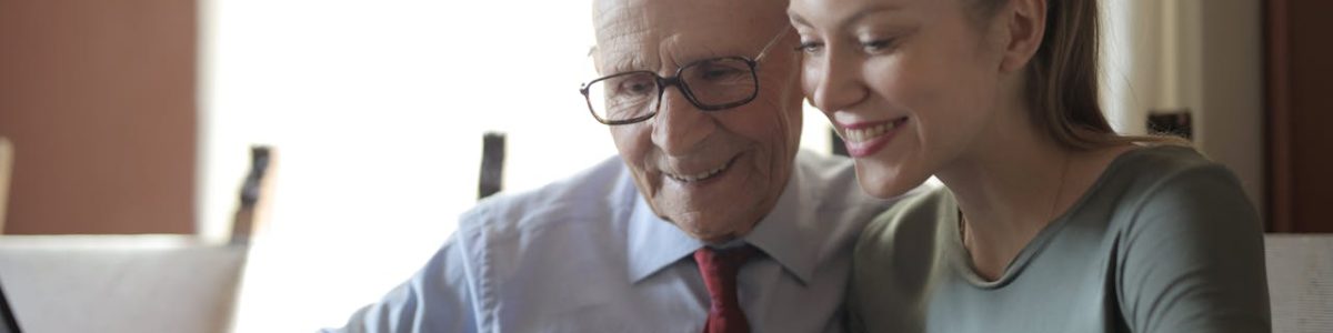 Smiling young woman in casual clothes showing smartphone to interested senior grandfather in formal shirt and eyeglasses while sitting at table near laptop